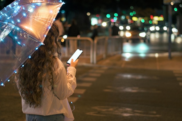 Mulher com um guarda-chuva com luzes e seu celular à noite