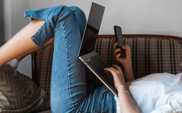 Mulher com um laptop está deitada em um sofá e usando um smartphone