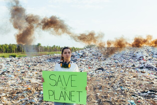 Foto mulher com um pôster salve o planeta, piquetes de ambiente contaminado com lixo, queimando fogo e fumaça negra