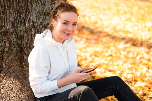 Mulher com um suéter branco com capuz está sentada no chão do parque e segurando um celular nas mãos.