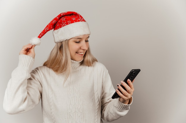 Mulher com um suéter branco e um chapéu de Papai Noel com um telefone nas mãos em um fundo cinza isolado