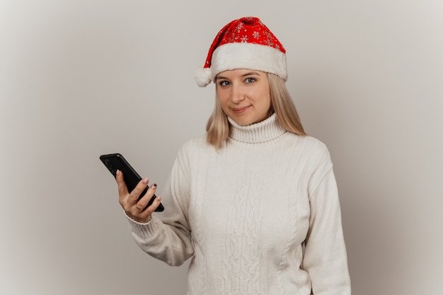 Mulher com um suéter branco e um chapéu de Papai Noel com um telefone nas mãos em um fundo cinza isolado