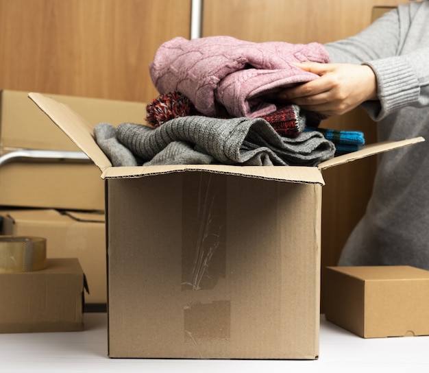 Foto mulher com um suéter cinza recolhe roupas em uma caixa, conceito de assistência e voluntariado, em movimento. atrás de uma pilha de caixas marrons