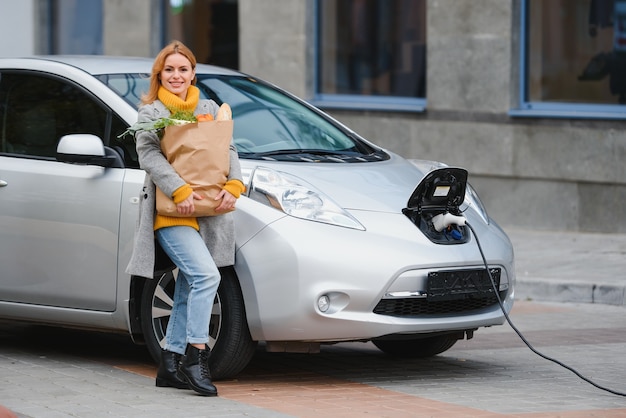 Foto mulher com um telefone celular perto de recarregar o carro elétrico. carregamento do veículo em uma estação de carregamento pública ao ar livre. conceito de compartilhamento de carro