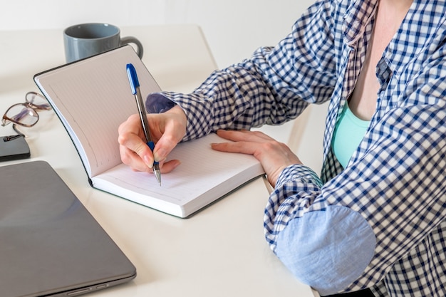 Mulher com uma camisa xadrez em sua mesa faz anotações em seu diário. Freelancer faz uma lista de tarefas pendentes.