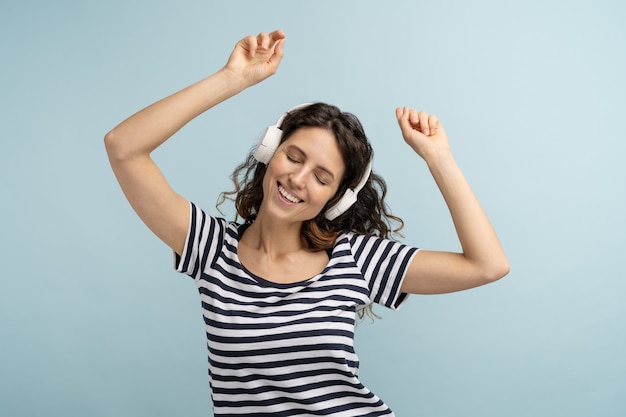 Mulher com uma camiseta listrada usa fones de ouvido sem fio, ouve música dançando com as mãos para cima