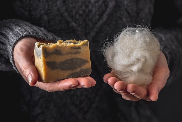 Mulher com uma camisola cinza quente segura um pouco de lã branca em uma mão e um pedaço de sabão de caxemira natural na outra mão