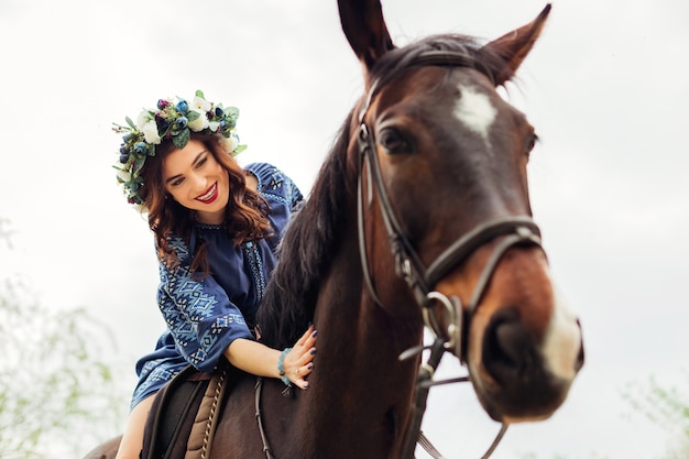 mulher com uma coroa na cabeça, sentada em um cavalo, sorrindo e acariciando o pescoço de um cavalo