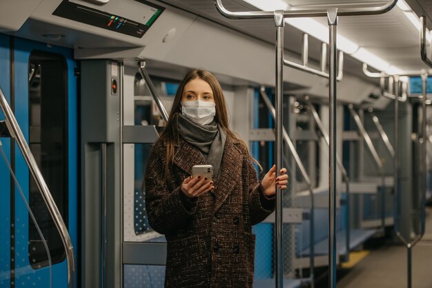 Mulher com uma máscara facial para evitar a propagação do coronavírus está de pé e segurando um smartphone em um vagão de metrô moderno. menina com máscara cirúrgica contra covid-19 está passeando em um trem do metrô.