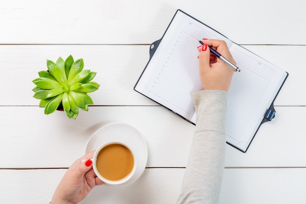 Mulher com unhas vermelhas segurando a xícara de café e escrevendo com uma caneta em um organizador de caderno perto de vaso na mesa de madeira branca.