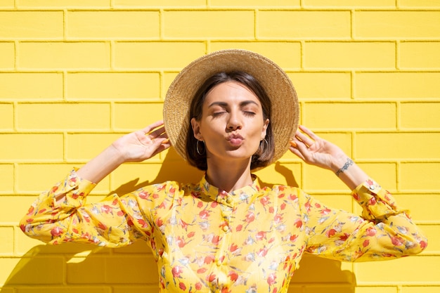 Mulher com vestido de verão amarelo e chapéu na parede de tijolo amarelo calma e positiva, gosta de dias ensolarados de verão