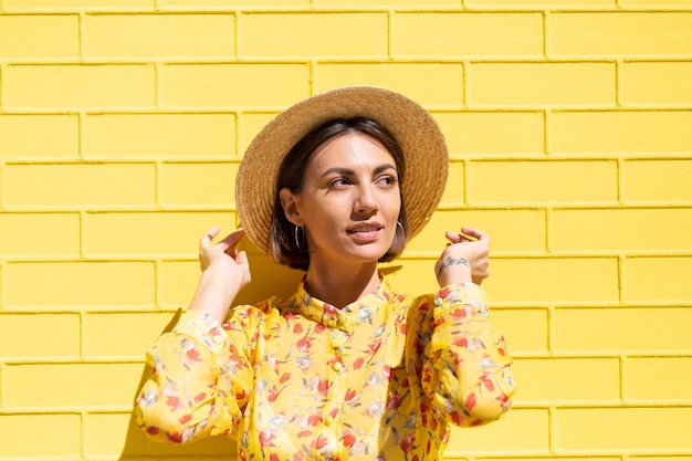 Foto mulher com vestido de verão amarelo e chapéu na parede de tijolo amarelo calmo e positivo