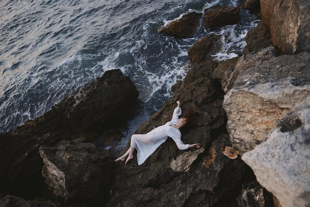 Mulher com vestido longo branco e cabelo molhado deitada em uma paisagem de penhasco rochoso