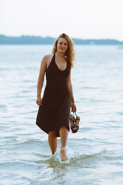 Mulher com vestido preto claro andando descalça à beira-mar na praia quente noite de verão