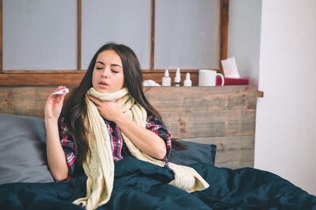 Mulher com vírus da gripe, deitada na cama, ela está medindo sua temperatura com um termômetro e tocando sua testa.