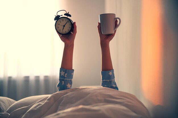 Mulher com xícara e despertador deitado na cama closeup hora da manhã