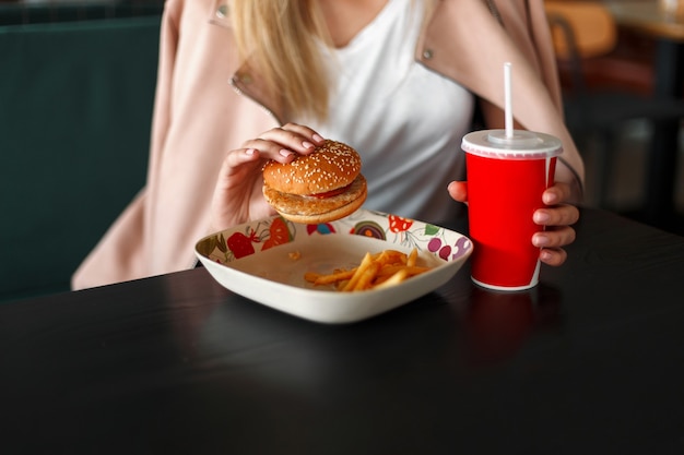 Mulher come fast food. Hambúrguer com batatas fritas e um copo vermelho com uma bebida na mesa de madeira