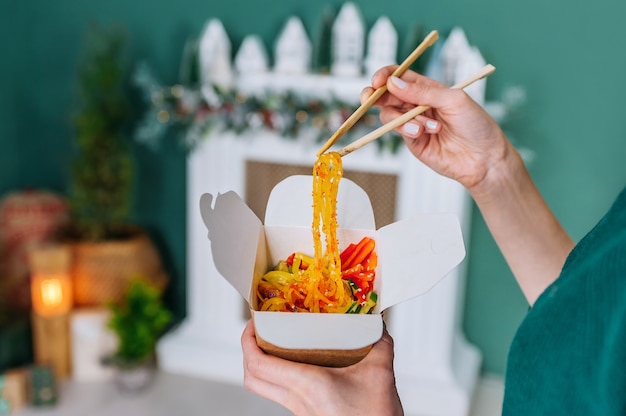 Foto mulher comendo macarrão wok de frutos do mar com os pauzinhos da caixa em fundo amarelo, closeup.