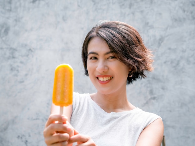 Mulher comendo picolés. Feliz linda mulher asiática cabelo curto vestindo casual camisa branca sem mangas segurando picolé amarelo no fundo da parede de concreto cinza no verão.