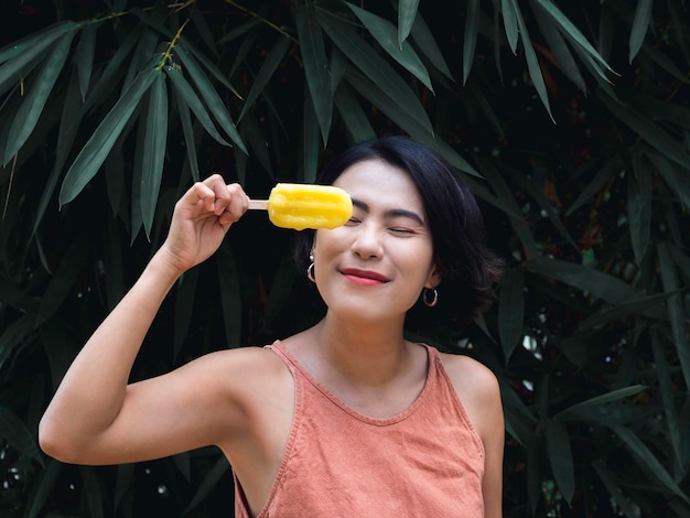 Mulher comendo picolés. Feliz linda mulher asiática vestindo blusa rosa casual segurando picolé amarelo sobre fundo de folhas de palmeira tropical verde, ao ar livre. Mulher sorridente, desfrutando de picolé no verão.