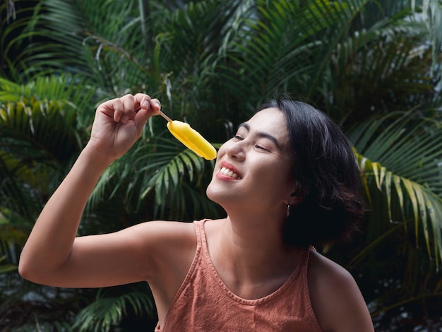 Mulher comendo picolés. Feliz linda mulher asiática vestindo blusa rosa casual segurando picolé amarelo sobre fundo de folhas de palmeira tropical verde, ao ar livre. Mulher sorridente, desfrutando de picolé no verão.