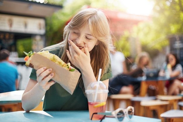 Mulher comendo taco sorrindo. mulher loira sardenta e faminta comendo junk food em uma praça de alimentação bebendo limonada