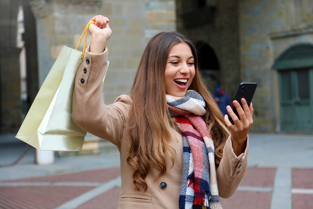 Mulher compradora feliz e animada rindo vendo o telefone levantado o braço com uma sacola de compras ao ar livre na rua da cidade