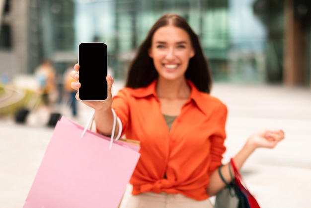 Mulher compradora segurando smartphone e sacolas de compras mostrando tela ao ar livre