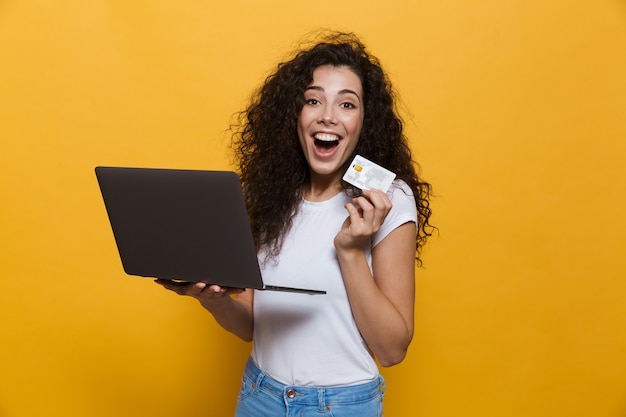 Foto mulher comprando 20 anos, vestindo roupas casuais, segurando um laptop preto e um cartão de crédito isolado no amarelo