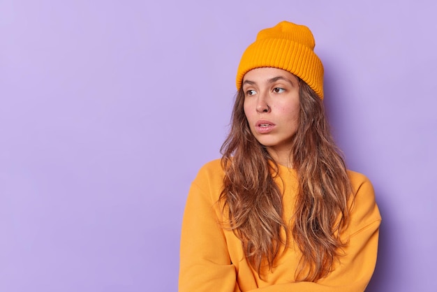 mulher concentrada, perdida em pensamentos, considera que algo está usando um chapéu laranja e um suéter posa em roxo com um espaço em branco