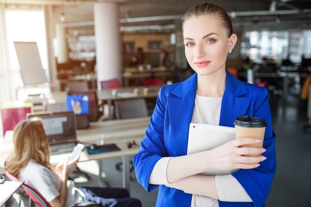 Mulher confiante e profissional está de pé no escritório e segurando uma xícara de café. Ela também tem um caderno nas mãos.