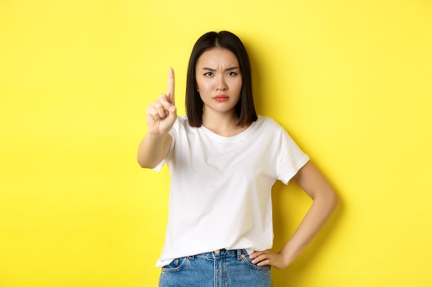 Mulher confiante e séria dizer não, mostrando o dedo estendido para parar e proibir algo ruim, franzindo a testa e olhando para a câmera autoconfiante, de pé sobre fundo amarelo.