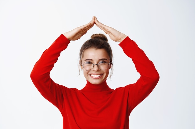 Mulher confiante em óculos mostrando as mãos do telhado, fazendo o telhado da casa e olhando determinada na frente, sorrindo satisfeita, parede branca