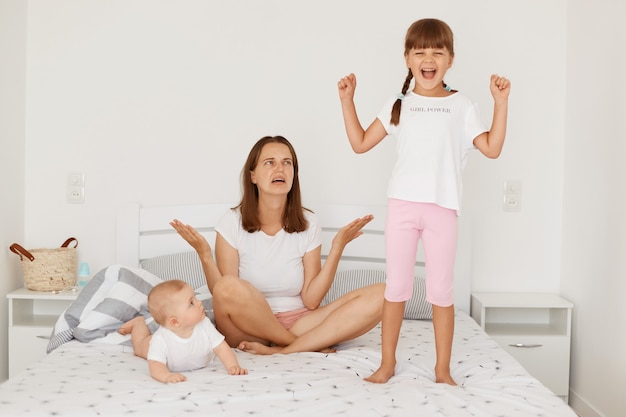 Mulher confusa e frustrada com cabelo escuro, sentada na cama com um bebê infantil e uma filha mais velha, levantando os braços, tendo uma expressão impotente, cansada de cuidar de crianças sozinha.