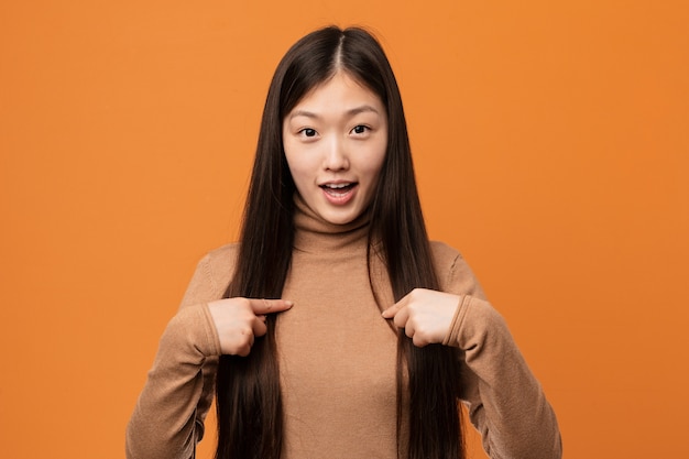 Mulher consideravelmente chinesa dos jovens surpreendida apontando dsi mesmo, sorrindo amplamente.