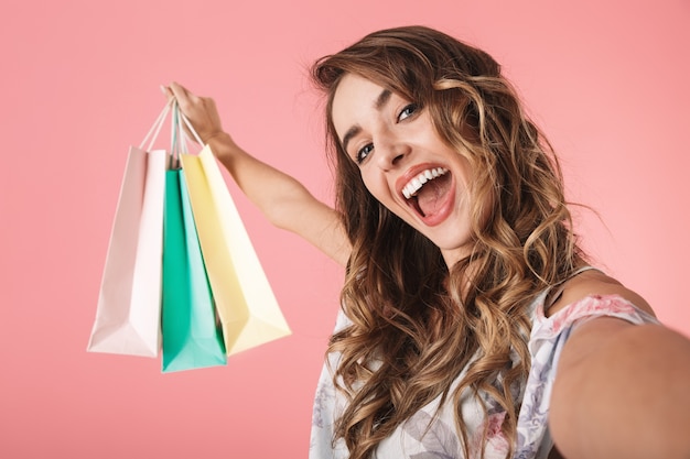 Mulher contente em vestido segurando sacolas de compras coloridas e tirando uma selfie, isolada em rosa