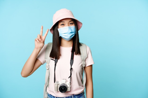 Mulher coreana feliz em câmera fotográfica de máscara facial médica mostrando vsigns de paz viajando durante o ...