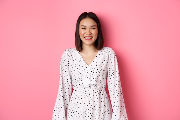 Mulher coreana feliz num vestido olhando para a câmera, sorrindo e rindo com uma expressão sincera, em pé sobre um fundo rosa