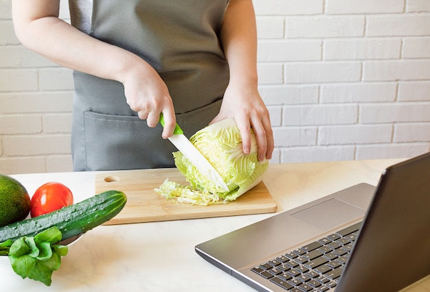 Mulher corta repolho na cozinha e usa o computador.