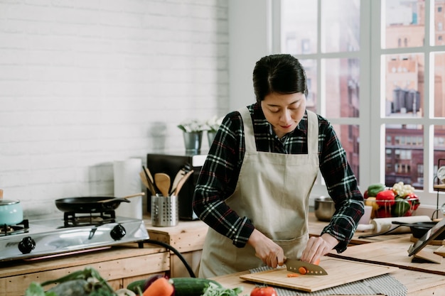 Mulher cortando cenoura na mesa de ilha de madeira na cozinha moderna em casa. Sorrindo senhora chinesa asiática assistindo programa de tv no tablet digital e cozinhando o jantar preparando uma refeição saudável com legumes frescos.