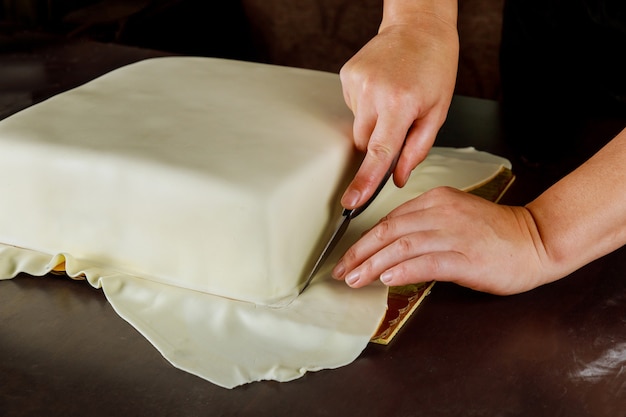 Mulher cortando fondant branco no bolo quadrado. Técnica de fazer bolo de casamento.