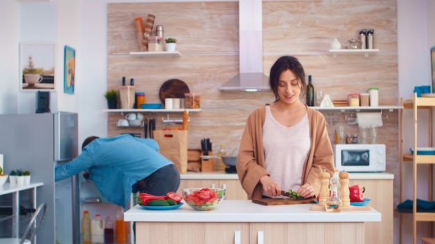 Mulher cortando pepinos na tábua de madeira para salada saudável. Marido abrindo geladeira. Cozinhar preparando alimentos orgânicos saudáveis, estilo de vida feliz juntos. Refeição alegre em família com vegetais