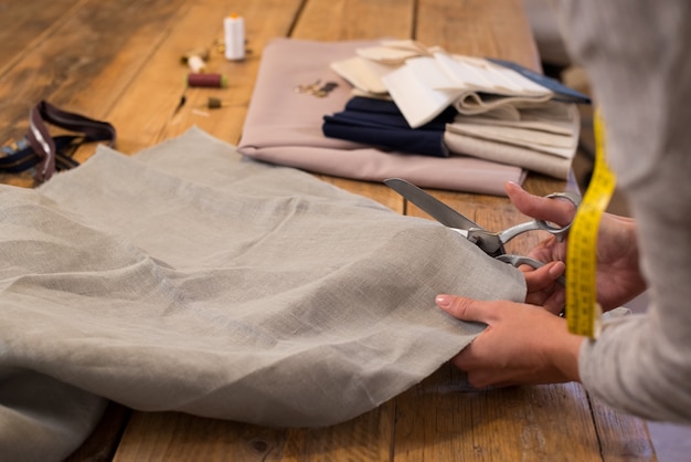 Foto mulher cortando um pedaço de pano na mesa no estúdio de moda. alfaiate de mulher trabalhando com tecido de tesoura na mesa. perto da costureira no trabalho, fazendo padrões de tecido.
