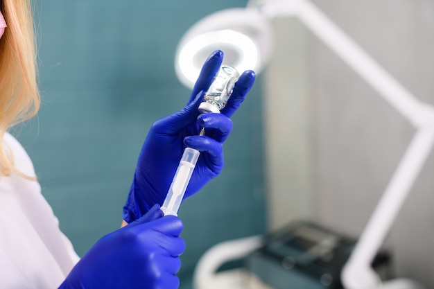 Mulher cosmetologista com as mãos usando luvas de borracha azuis e encher a seringa com o medicamento da ampola