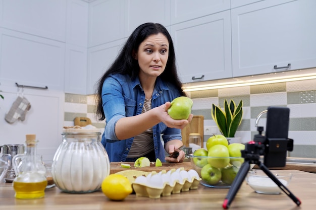 Mulher cozinhando em casa na cozinha falando online usando videochamada no smartphone