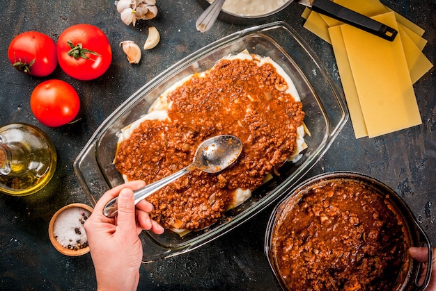 Mulher cozinhando lasanha clássica caseira à bolonhesa, na mesa azul escura; com ingredientes, vista superior copyspace, mãos na foto