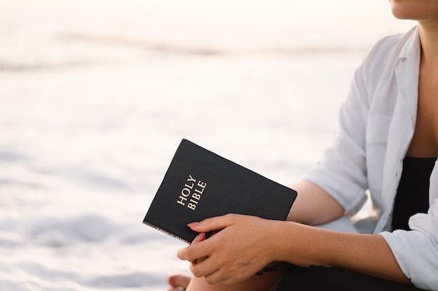 Mulher cristã segura a Bíblia nas mãos, lendo a Bíblia Sagrada no mar durante o belo pôr do sol