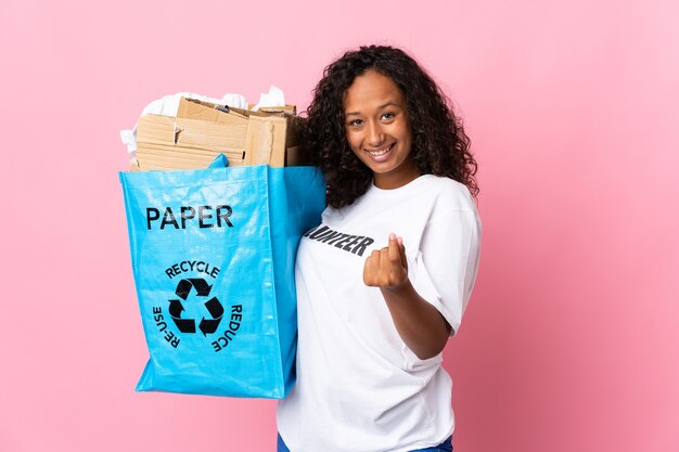 Mulher cubana segurando um saco de reciclagem cheio de papel para reciclar isolado em rosa fazendo gesto de dinheiro