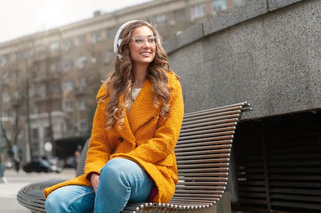 Mulher Óculos Ouvir Música Fone de Ouvido Banco de Assento Ao Ar Livre Vestido Elegante Elegante Casaco Amarelo Sorriso Caucasiana Mulher 30 anos Desfrute de Podcast ou Audiobooks lá fora