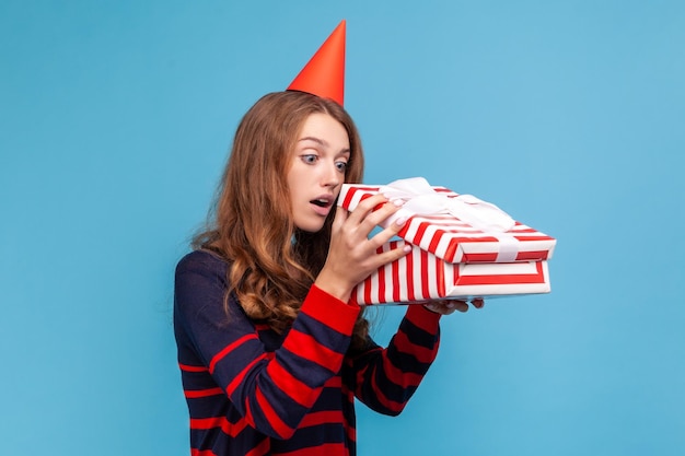 Mulher curiosa com cabelos ondulados vestindo suéter listrado estilo casual e cone de festa segurando a caixa de presente olhe para dentro com interesse o que está dentro do estúdio interior isolado em fundo azul